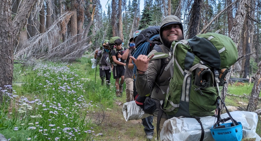 A group of backpackers hike through a wooded area. The person in front smiles and gives the camera a hang loose sign. 